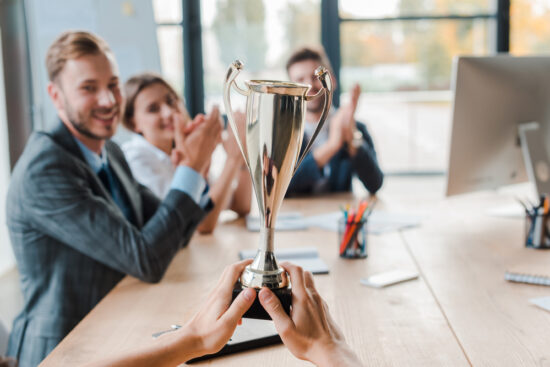 Hands holding a trophy in an office setting while colleagues applaud