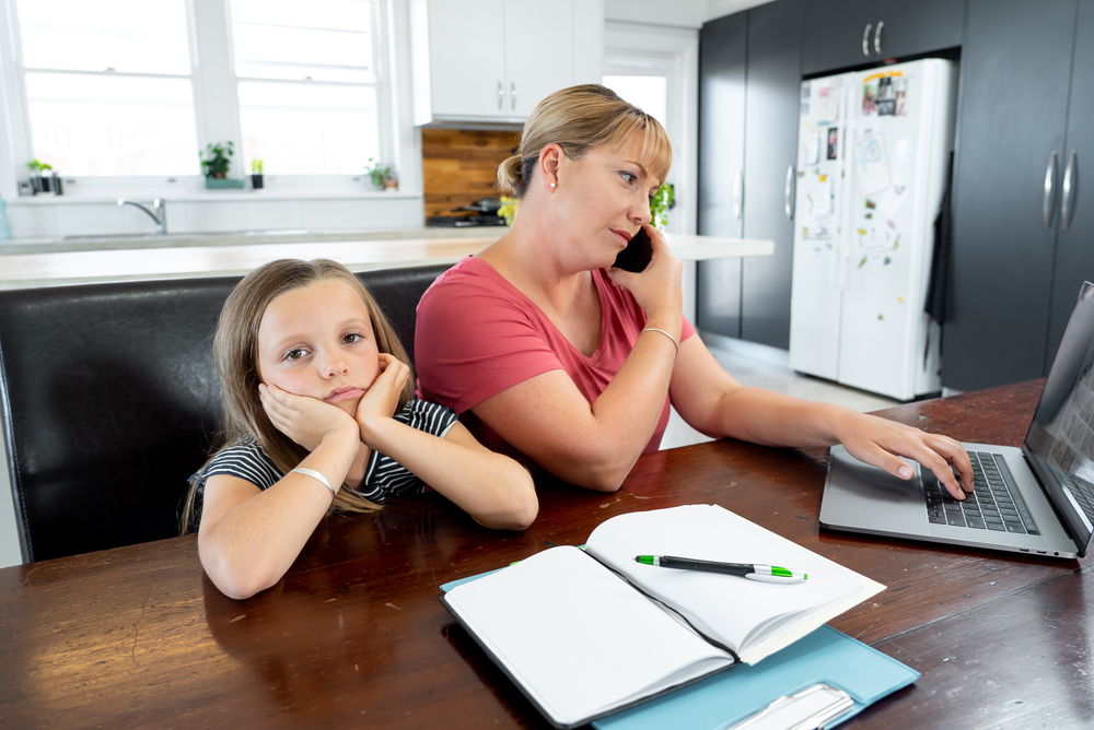 a busy mom is on the phone and looking at her laptop at the kitchen table while her daughter waits for help with homework