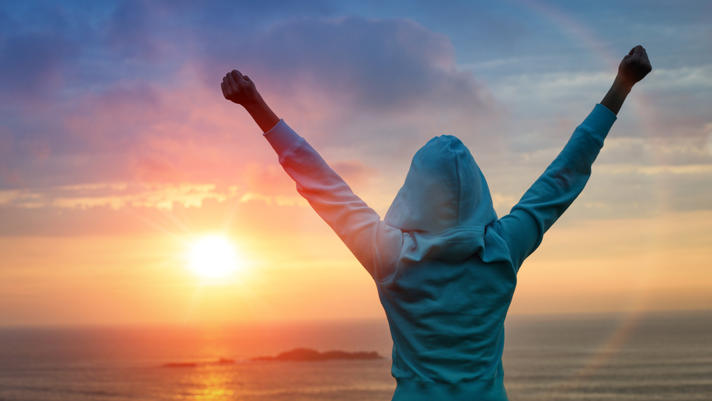 A woman in a hoodie with her arms outstretched in the sunset.