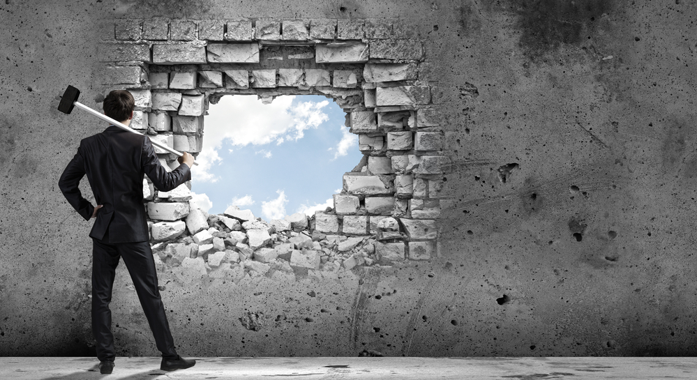 A business professional with a sledge hammer looks through an opening in a wall where you see blue sky