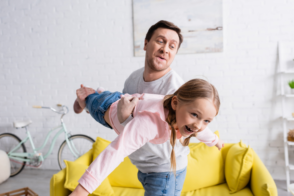 A dad is holding his daughter to let her fly through the living room.