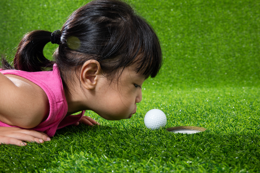 Asian little girl lying on grass and blowing the ball into a hole