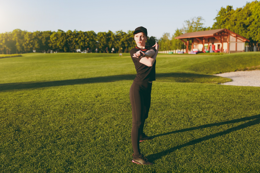 Young athletic girl in black uniform, cap doing sport exercises, warm-up, stretching before running on green lawn in golf course outdoors on sunny summer day.