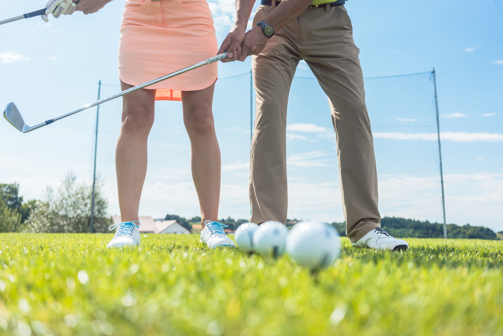Low section of man and woman holding iron clubs, while practicing together the correct grip and move for playing golf on the green grass of a professional ground