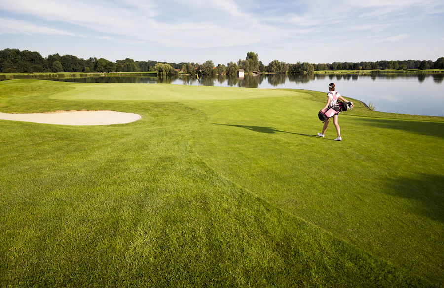 Female golf player walking towards green on fairway carrying golf bag with clubs, lake in background.